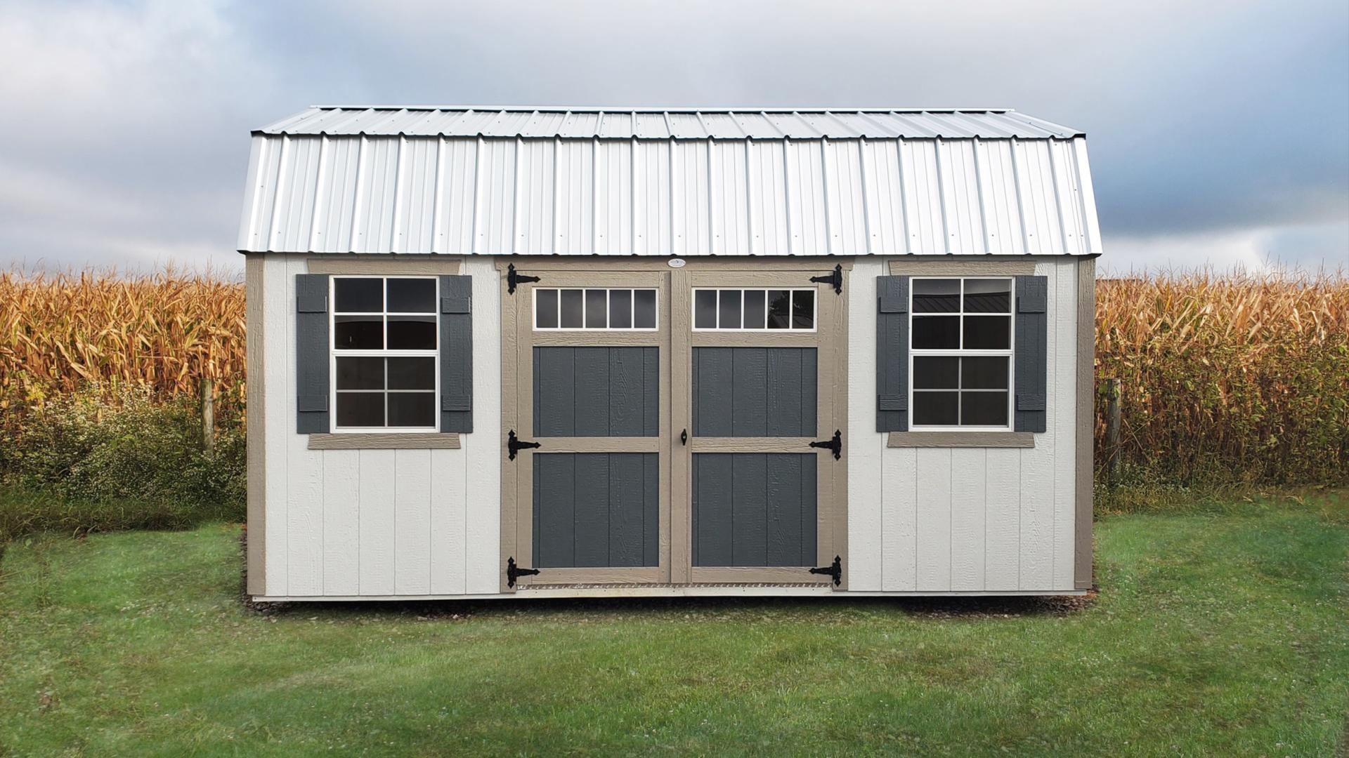 Compass High Barn shed with white siding, tan trim, white metal roofing, blue double doors, and 2 windows with blue shutters sitting in a backyard in front of a cornfield.