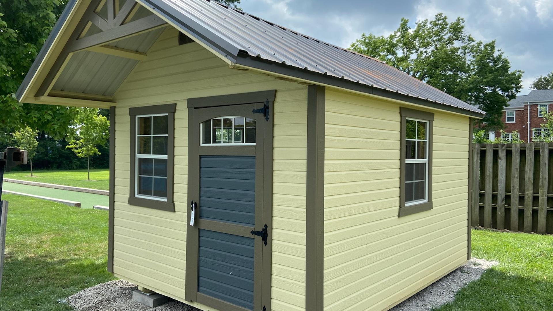 Yellow garden shed with green trim, a blue single door, multiple windows, door awning, and a metal roof that sits in a backyard and can be used as a kid's playhouse.