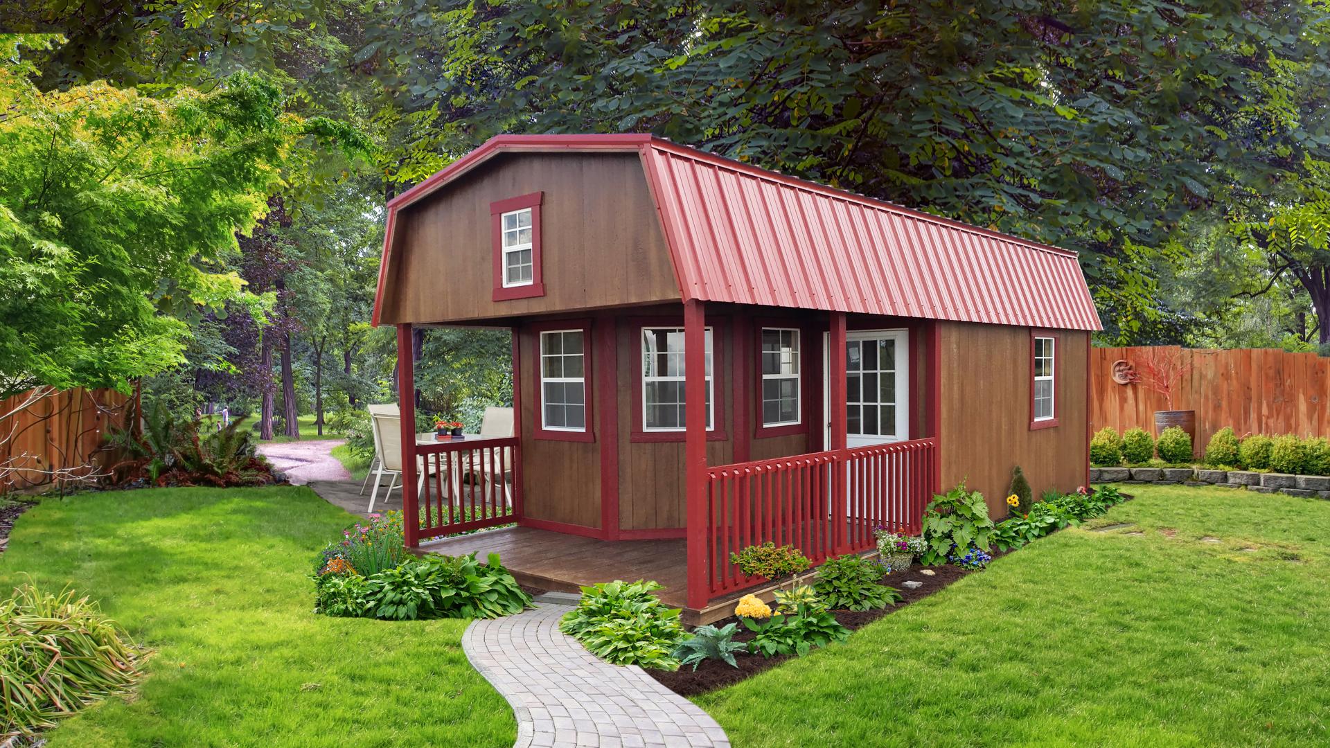 Brown lofted cabin with red trim, red metal roof, front porch, front doors, and many windows sitting in a residential area with a walkway and fence in the background. 