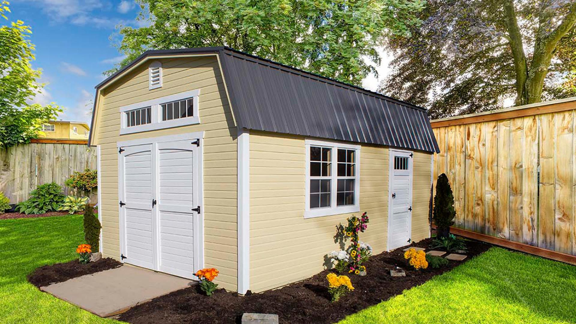 High barn shed with light yellow siding, white trim, white double doors, windows, a side door entrance, and a dark gray metal roof sitting in a fenced in backyard.