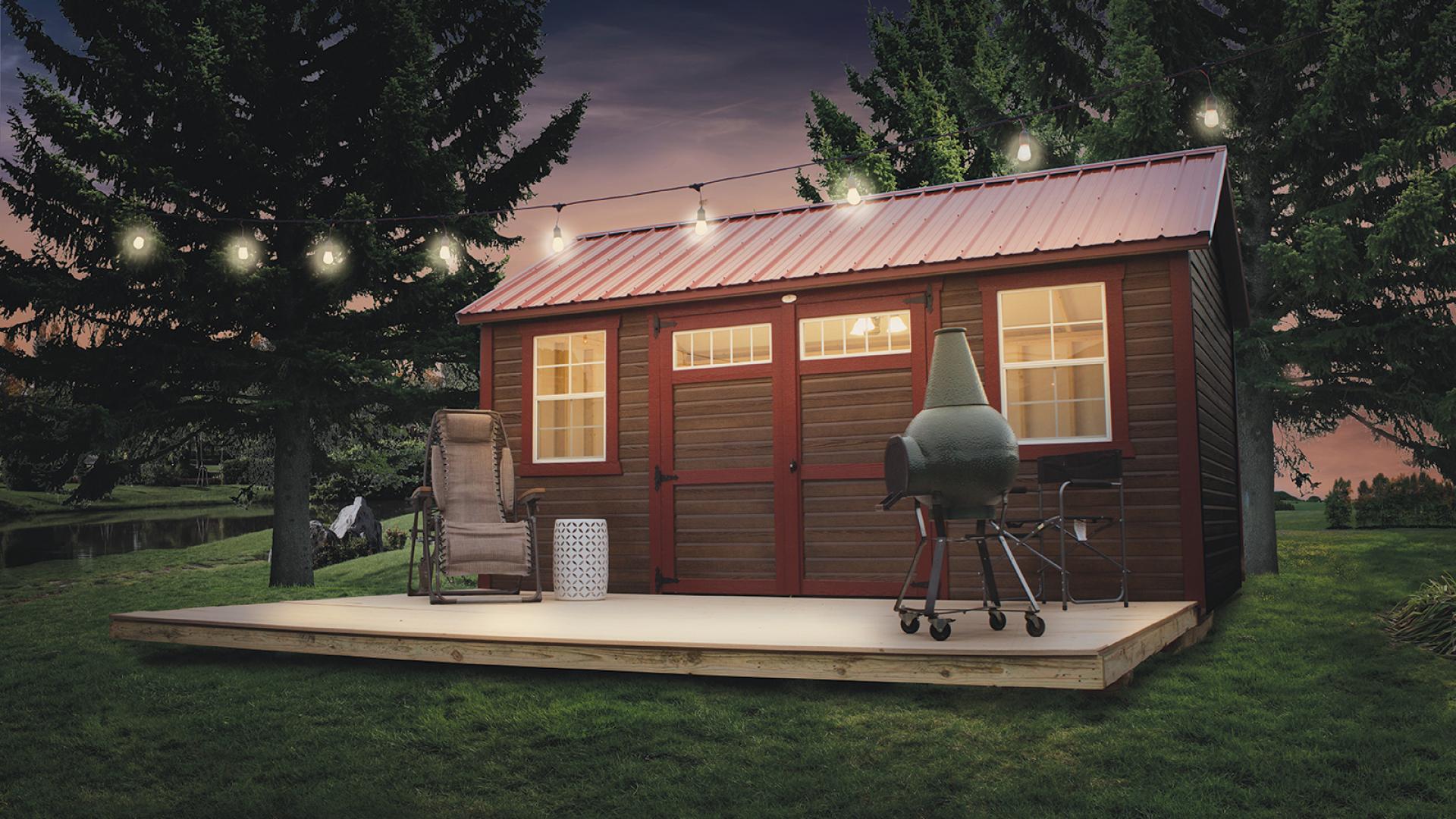 Tiny house made from a garden shed with brown siding, red trim, and red roofing sitting by a river with a porch that has hanging lights, a grill, and outdoor seating.